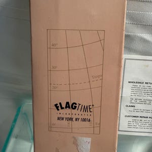 May include: A rectangular cardboard box with a light brown exterior. The box features a grid pattern with degree markings and the text "FLAGTIME INCORPORATED NEW YORK, NY 10016". The bottom of the box reads "MADE IN HONG KONG".