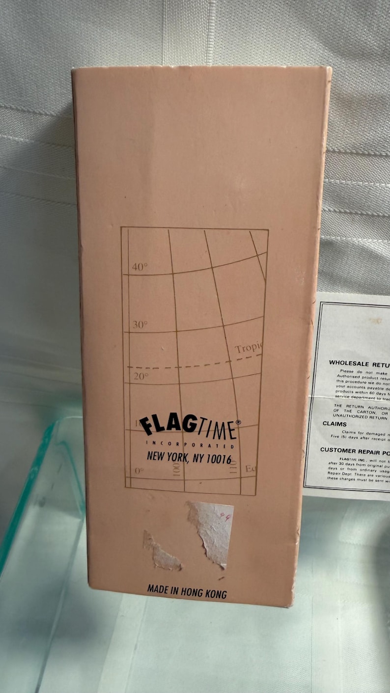 May include: A rectangular cardboard box with a light brown exterior. The box features a grid pattern with degree markings and the text "FLAGTIME INCORPORATED NEW YORK, NY 10016". The bottom of the box reads "MADE IN HONG KONG".