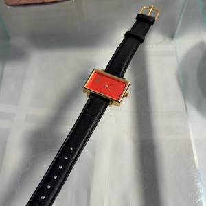 May include: A rectangular gold-toned watch with a red face and black leather band. The watch face has a small logo. The watch is displayed on a clear surface. The watch is from the brand "FLAGTIME".
