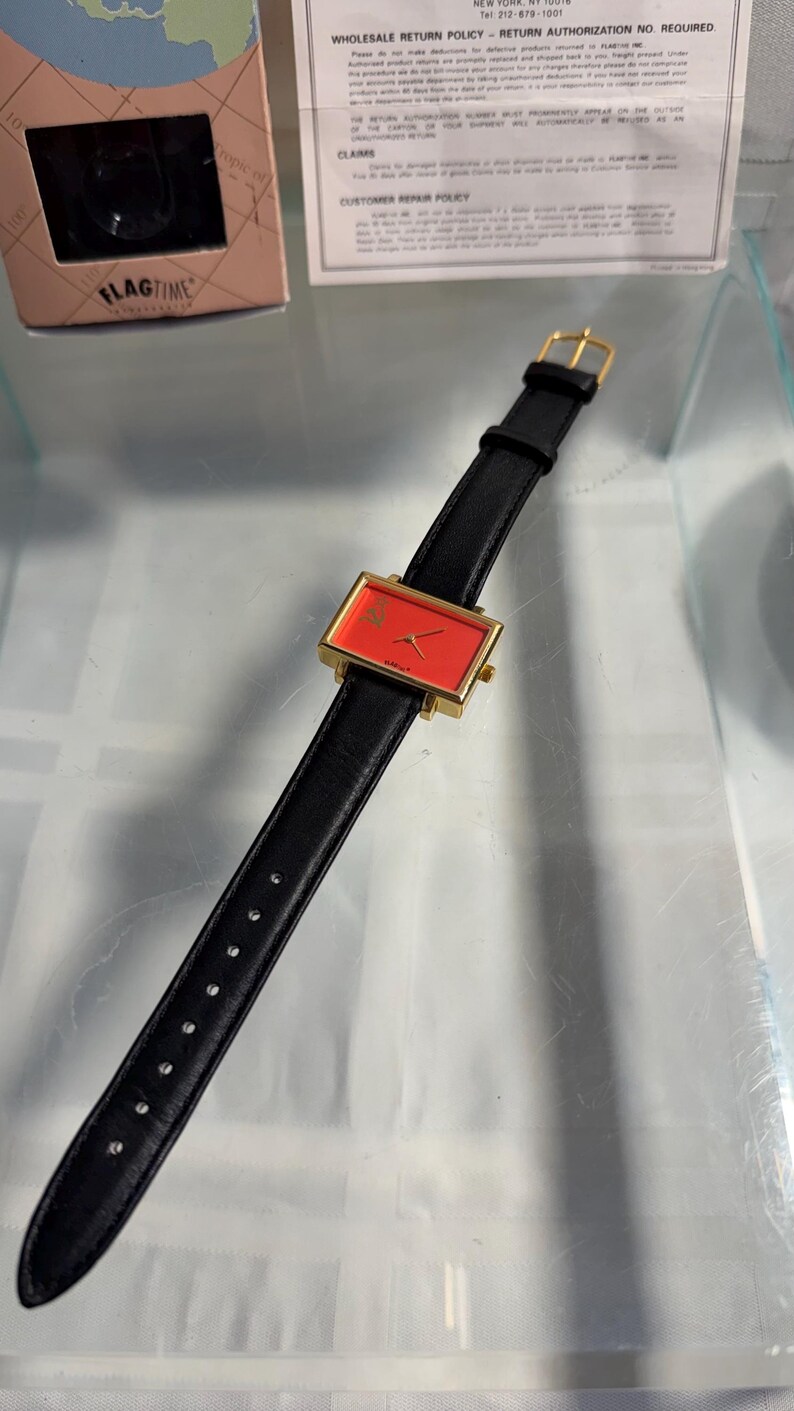 May include: A rectangular gold-toned watch with a red face and black leather band. The watch face has a small logo. The watch is displayed on a clear surface. The watch is from the brand "FLAGTIME".