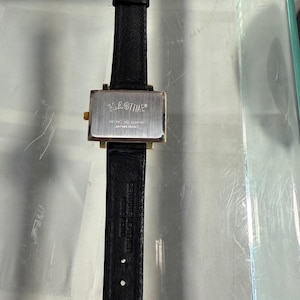 May include: A rectangular wristwatch with a black leather band and a gold-colored buckle. The watch face is silver with the brand name "FLAGTIME" printed on it. The watch is displayed on a clear surface.
