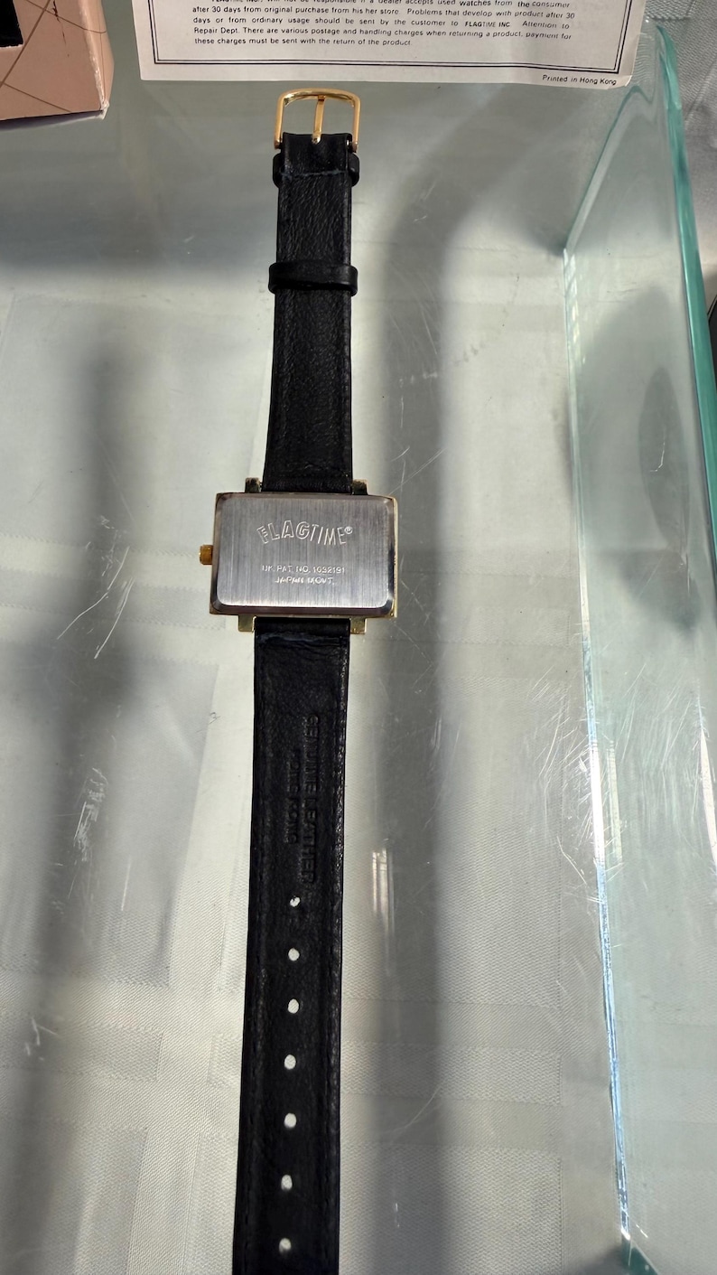 May include: A rectangular wristwatch with a black leather band and a gold-colored buckle. The watch face is silver with the brand name "FLAGTIME" printed on it. The watch is displayed on a clear surface.