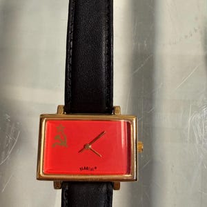 May include: A rectangular wristwatch with a gold-colored case and a red face. The watch features a black leather band. The watch face has a gold-colored emblem and the word "FLAGTIME".