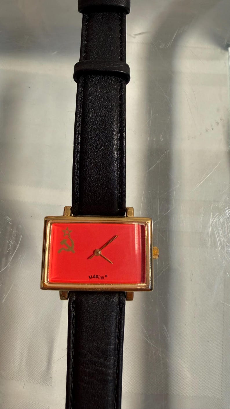 May include: A rectangular wristwatch with a gold-colored case and a red face. The watch features a black leather band. The watch face has a gold-colored emblem and the word "FLAGTIME".