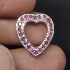 Pode incluir: Um pingente em forma de coração feito de metal prateado, com uma borda cravejada com numerosas pequenas pedras preciosas redondas de um vibrante tom vermelho rosado. O centro do coração é aberto, criando um design de moldura oca.