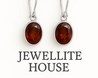 Puede incluir: Un par de pendientes plateados con piedras preciosas ovaladas, facetadas, de color marrón rojizo. Los pendientes tienen un diseño de gancho simple. El texto "JEWELLITE HOUSE" se muestra debajo de los pendientes.