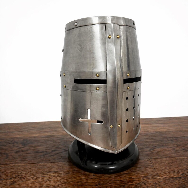 May include: A silver-colored, medieval-style helmet with a cross-shaped opening and eye slits. The helmet is riveted and sits on a black base. The metal has a slightly weathered appearance.