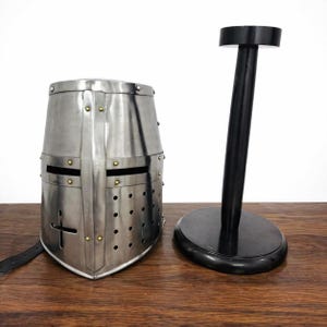 May include: A silver-colored medieval crusader helmet with a cross design, rivets, and eye slits. A black helmet stand is next to the helmet. The helmet is made of metal and has a leather strap.