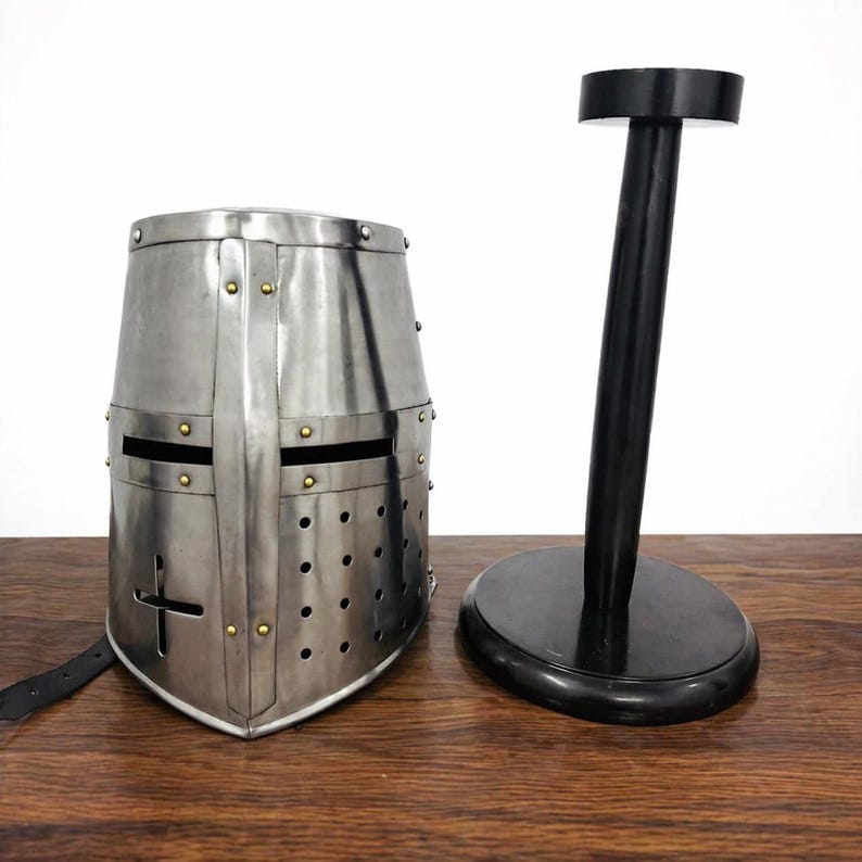 May include: A silver-colored medieval crusader helmet with a cross design, rivets, and eye slits. A black helmet stand is next to the helmet. The helmet is made of metal and has a leather strap.