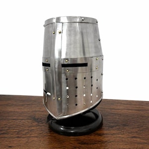 May include: A silver-colored, medieval-style helmet with a cross design and rivet details. The helmet has eye slits and ventilation holes. It is displayed on a black, rounded base, and is made of metal.