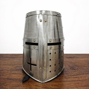 May include: A silver-colored, medieval-style helmet with a cross design. The helmet has a rounded top, horizontal eye slits, and small ventilation holes. Rivets secure the metal plates. The helmet is made of steel and has a polished finish.