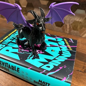 May include: A black goat figurine with purple wings and red eyes sits atop a book titled "This Inevitable Ruin." The book has a black cover with turquoise and pink text. The figurine is a detailed, three-dimensional model.