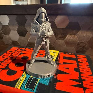 May include: A gray figurine of a person in a hooded jacket and suit, standing on a round base. The base has the name "Clitch" on it. The figurine is on top of a book with the title "The Dungeon Anarchist's Guide" and the author's name "Matt Dinniman".