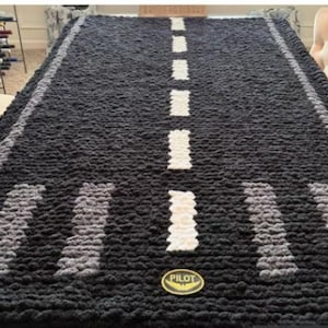 May include: A large, rectangular, black and gray crocheted blanket with a road design. The blanket features a white dotted center line and gray and white runway markings. A gold-colored "PILOT" emblem is visible.
