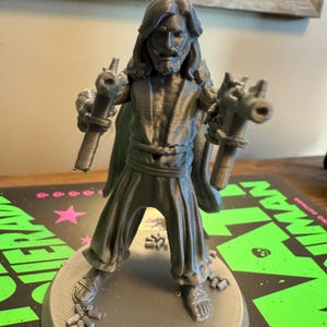 Pu&ograve; includere: Figurina grigia stampata in 3D di un uomo con capelli lunghi e barba, che tiene in mano due oggetti simili a pistole. La figura indossa una veste e sandali, in piedi su una base circolare. La figurina &egrave; alta circa 15 cm.