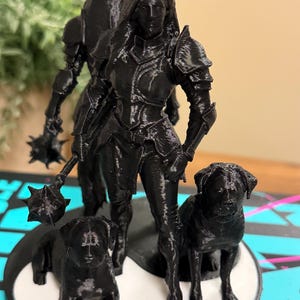 May include: A black 3D-printed figurine of a warrior woman in armor, standing with two dogs. The warrior holds a spiked mace. The dogs are positioned at the base of the figurine. The figurine is on a round base.