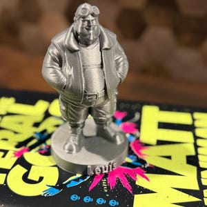 May include: A silver-colored figurine of a man wearing a bomber jacket, goggles, and boots. The figurine is standing on a round base with the name "Louis" on it. The figurine is placed on top of a book with the title "State of the Feral Man".