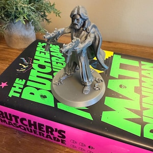 Pu&ograve; includere: Figurina grigia stampata in 3D di una persona che tiene due pistole, su una base circolare. La figurina &egrave; posizionata su un libro intitolato "The Butcher's Masquerade" con copertina nera e testo verde neon e rosa.