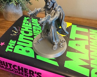 May include: A gray 3D-printed figurine of a person holding two guns, standing on a circular base. The figurine is placed on a book titled "The Butcher's Masquerade" with a black cover and neon green and pink text.