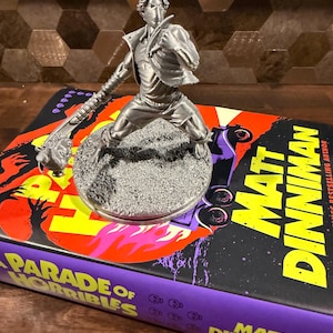 May include: A silver-colored figurine of a character in a dynamic pose, wearing a mask and holding a spiked weapon, standing on a circular base. The figurine is placed on a book titled Parade of Horribles by Matt Dinniman with a black cover.
