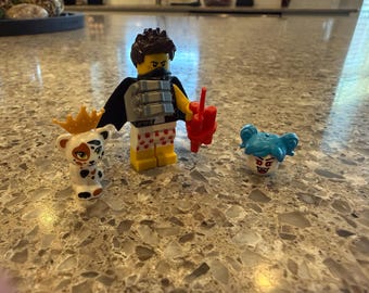 Può includere: Una minifigure in plastica con capelli castano scuro, mantello nero e pantaloncini bianchi con impronte di zampe rosse, che tiene in mano un accessorio rosso. Accanto si trova un piccolo animale bianco con corona dorata e una testa blu con zanne.