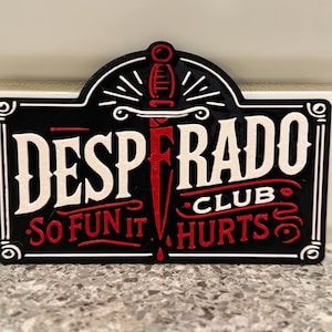 May include: Black and white rectangular sign with the words "DESPERADO CLUB" and "SO FUN IT HURTS" in a vintage style. A red dagger is in the center of the sign.