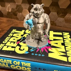 May include: A silver-colored figurine of a muscular bear holding a blue object, standing on a round base. The figurine is placed on top of a book titled "The Gate of the Feral Gods" by Matt Dinniman.