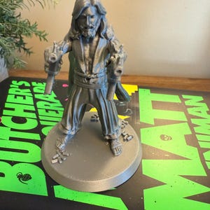 Pu&ograve; includere: Una statuetta grigia stampata in 3D di un uomo con capelli lunghi e barba, che tiene due pistole. La figura &egrave; su una base circolare. La base &egrave; su un libro con testo verde e nero.