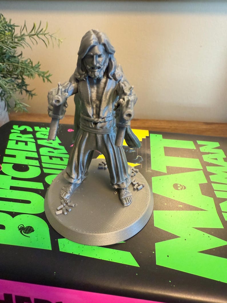 Pu&ograve; includere: Una statuetta grigia stampata in 3D di un uomo con capelli lunghi e barba, che tiene due pistole. La figura &egrave; su una base circolare. La base &egrave; su un libro con testo verde e nero.