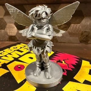 May include: A silver figurine of a fairy with crossed arms and an angry expression. The fairy has large wings and is standing on a round base. The figurine is placed on top of a book with the words "Dungeon Crawl" in yellow.