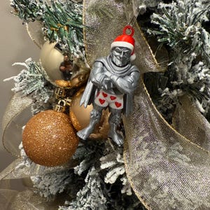 May include: A silver-colored Christmas ornament of a character wearing a Santa hat and heart-patterned underwear. The ornament is hanging on a decorated Christmas tree with gold ornaments and ribbon.