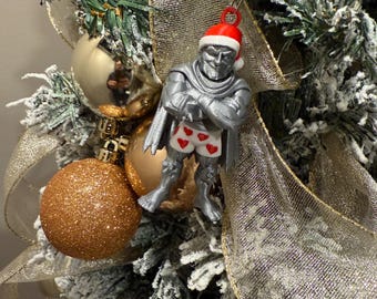 Può includere: Un ornamento natalizio color argento raffigurante un personaggio con un cappello da Babbo Natale e mutande a forma di cuore. L'ornamento è appeso a un albero di Natale decorato con palline e nastri dorati.