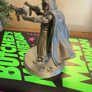 Pu&ograve; includere: Figurina grigia stampata in 3D di un personaggio con due pistole, un mantello e capelli lunghi. La figurina &egrave; su una base circolare. La base &egrave; su un libro con il testo "BUTCHER'S RAGE" e "THE BOYS" in verde e rosa.
