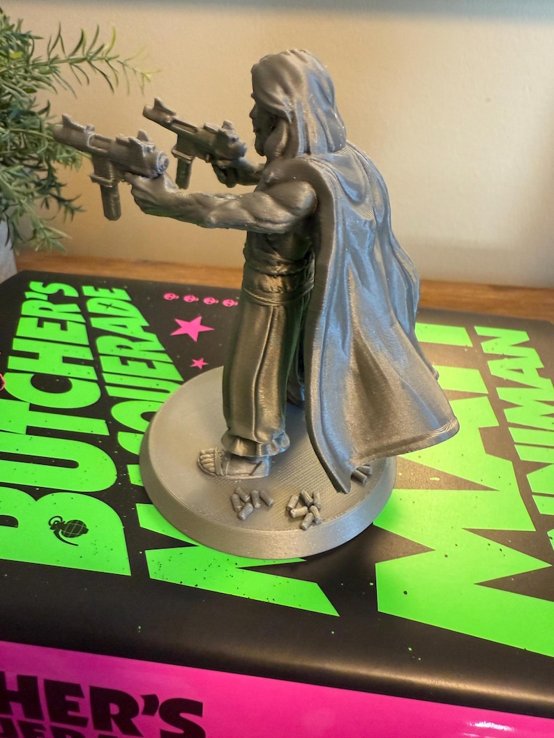 Pu&ograve; includere: Figurina grigia stampata in 3D di un personaggio con due pistole, un mantello e capelli lunghi. La figurina &egrave; su una base circolare. La base &egrave; su un libro con il testo "BUTCHER'S RAGE" e "THE BOYS" in verde e rosa.