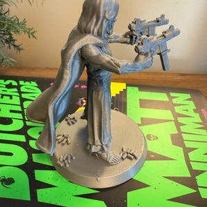 Pu&ograve; includere: Figurina grigia stampata in 3D di una figura incappucciata che tiene due pistole. La figura &egrave; su una base circolare con piccoli dettagli. La base poggia su un libro con testo verde e nero.