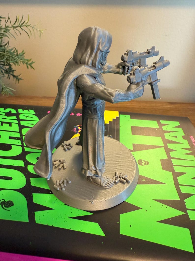 Pu&ograve; includere: Figurina grigia stampata in 3D di una figura incappucciata che tiene due pistole. La figura &egrave; su una base circolare con piccoli dettagli. La base poggia su un libro con testo verde e nero.