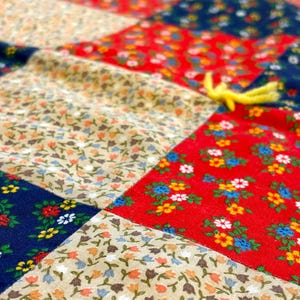 May include: Close-up of a patchwork quilt with a variety of floral patterns. The quilt features red, blue, and beige fabric squares, each adorned with small, colorful flowers. Yellow yarn accents are visible.