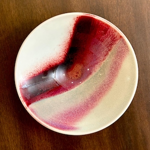 May include: A round ceramic plate with a cream-colored base and a vibrant abstract design. The design features a bold splash of deep red and black, transitioning into lighter shades of pink and gray. The plate has a glossy finish.