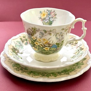 May include: A white porcelain teacup and saucer set featuring a scalloped edge and a detailed illustration of a small mouse nestled among yellow primrose flowers and green foliage. The interior of the cup displays a small floral bouquet design.