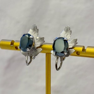 May include: A pair of silver-toned clip-on earrings featuring a textured leaf design. Each earring is centered with a single, oval-shaped, dark blue faceted gemstone held in place by four prongs, creating a botanical-inspired accessory.
