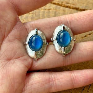 May include: A pair of vintage silver-toned cufflinks. Each cufflink features an oval, textured silver frame with a vibrant blue, rounded center. The cufflinks are displayed against a neutral background.