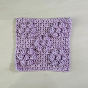 May include: A square, lavender crochet piece featuring a textured floral pattern. The flowers are created with raised stitches, adding dimension to the soft, handmade textile. The piece is likely a coaster or a small decorative item.