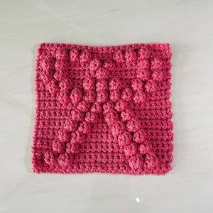May include: A square, handmade crochet item in a vibrant shade of pink. The piece features a textured design with raised, rounded bobbles arranged in a repeating pattern. The stitches are tightly woven, creating a dense and visually appealing texture.