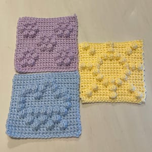 Puede incluir: Tres cuadrados de ganchillo en colores pastel: lavanda, azul claro y amarillo. Cada cuadrado presenta un patrón texturizado y en relieve de bolitas. Los cuadrados están hechos a mano, probablemente para un proyecto de artesanía.