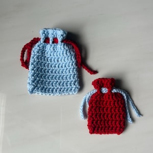 May include: Two small, handmade crochet drawstring bags. One is light blue with red drawstring, the other is red with light blue drawstring. The bags are likely for small gifts or trinkets.