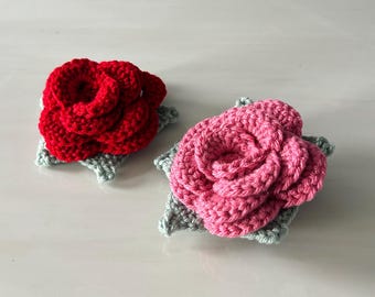 May include: Two handmade crochet rose flowers. One is bright red, the other is pink. Each rose is attached to a light green, star-shaped base. The roses are made of soft yarn and are likely used as decorative accents.