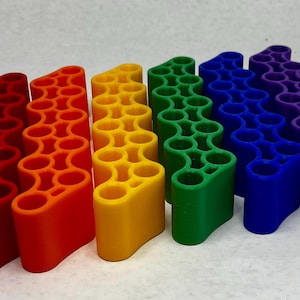 May include: A set of six colorful, wavy-shaped organizers in a rainbow of colors: red, orange, yellow, green, blue, and purple. Each organizer has multiple cylindrical holes. The objects are arranged in a row, showcasing a vibrant spectrum.