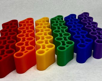 May include: A set of six colorful, wavy-shaped organizers in a rainbow of colors: red, orange, yellow, green, blue, and purple. Each organizer has multiple cylindrical holes. The objects are arranged in a row, showcasing a vibrant spectrum.