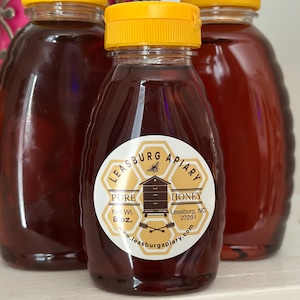 Puede incluir: Tres botellas de vidrio de miel con tapas amarillas. La botella central tiene una etiqueta que dice "Leasburg Apiary Pure Honey" y "Net Wt. 227g". La miel es de un rico color &aacute;mbar.
