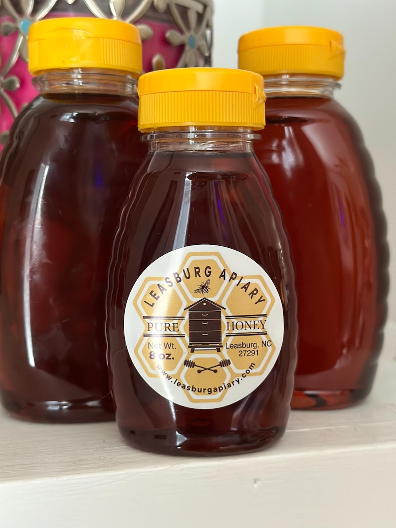 Puede incluir: Tres botellas de vidrio de miel con tapas amarillas. La botella central tiene una etiqueta que dice "Leasburg Apiary Pure Honey" y "Net Wt. 227g". La miel es de un rico color &aacute;mbar.